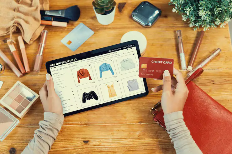 Top-down view of a consumer's hands holding a red credit card and a tablet displaying apparel, representing Amazon Product Listing Services(FAQs) by Brandock on a wooden desk with cosmetics.