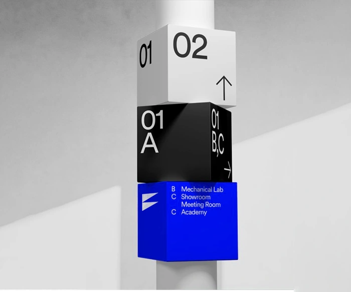 A modern geometric totem display illustrating a business growth concept titled While You Wait, Competitors Are Already Scaling by Brandock. The image features three large blocks stacked vertically on a white central pillar. The top block is white with black numbers "01" and "02" and an upward arrow. The middle block is black with white markings including "01 A" and "01 B, C" with a right-pointing arrow. The bottom block is a vibrant blue showing a white abstract wing-shaped logo on the left and white text labels for corporate directories including "B Mechanical Lab," "C Showroom Meeting Room," and "C Academy" on a clean grey background.