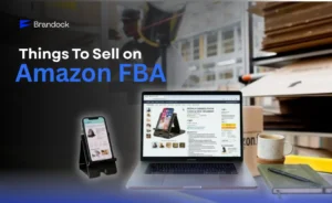A promotional e-commerce banner exploring Things to Sell on Amazon FBA by Brandock. The top-left corner features the bold title text and the blue corporate logo. In the center, an open laptop displays an active Amazon product listing page for a phone stand, placed next to a physical smartphone resting on a black desk organizer stand. The desktop setup is completed with a ceramic coffee mug, a green notebook with a pen, and a blurred background showcasing an e-commerce fulfillment warehouse with cardboard shipping boxes.