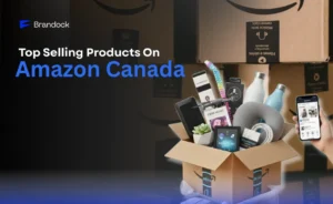 A promotional e-commerce banner showcasing the Top Selling Products on Amazon Canada by Brandock. On the left side, against a dark, shadowed backdrop of stacked cardboard shipping boxes, the bold headline text is displayed with "Amazon Canada" highlighted in bright blue, sitting below the blue corporate Brandock logo. On the right side, an open cardboard box taped with the Amazon smile logo is overflowing with various retail items, including a small succulent plant, e-readers, a neck pillow, water bottles, and a drone. A hand holds a smartphone next to the box, displaying an active product listing page on the screen.